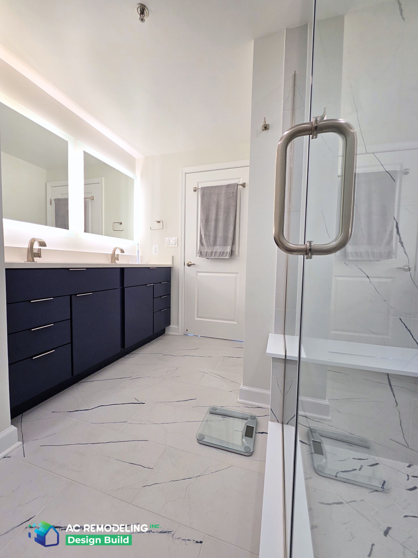 Wide view of the navy double vanity, backlit mirrors and glass shower entry